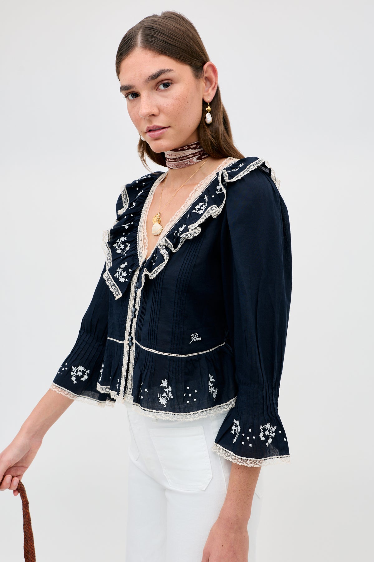 A woman with straight brown hair wears the Solene - Flower Trail Embroidery Navy blouse, styled with white pants, gold jewelry, and a brown ribbon choker, standing against a plain light gray background.