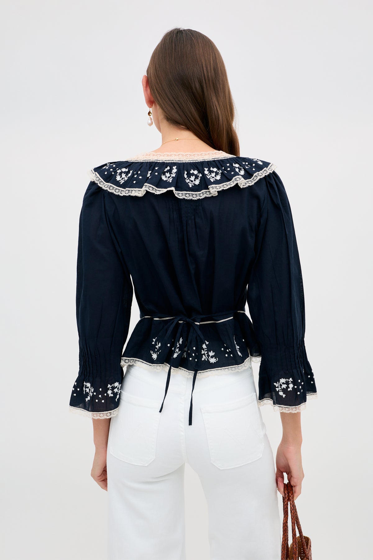 A woman with long brown hair wears the Solene - Flower Trail Embroidery Navy blouse and white high-waisted pants, holding a brown bag against a plain light background.