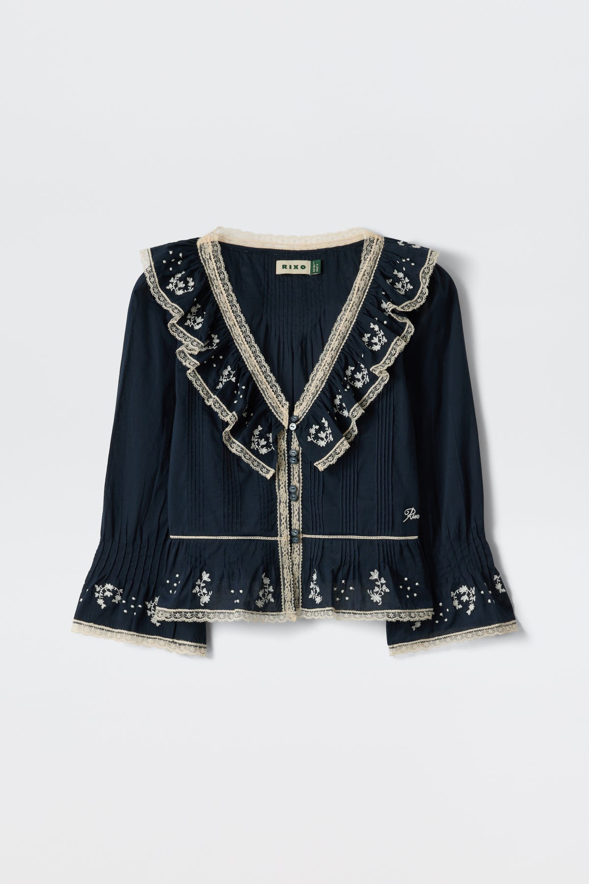The Solene - Flower Trail Embroidery Navy blouse is a long-sleeved vintage top in navy with cream lace trim, ruffled collar and chest, and white floral embroidery on the sleeves, hem, and yoke, shown against a white background.