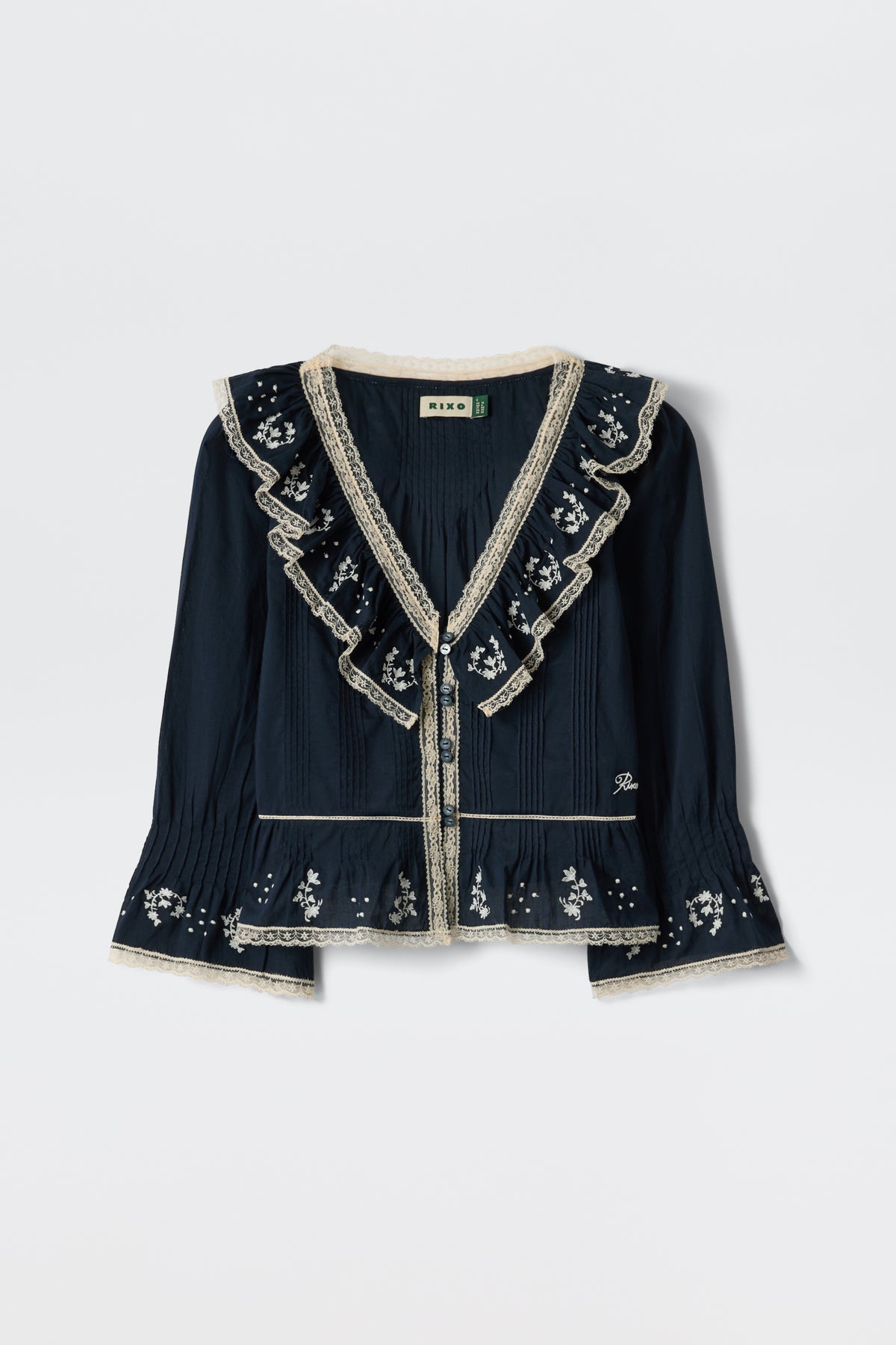The Solene - Flower Trail Embroidery Navy blouse is a long-sleeved vintage top in navy with cream lace trim, ruffled collar and chest, and white floral embroidery on the sleeves, hem, and yoke, shown against a white background.