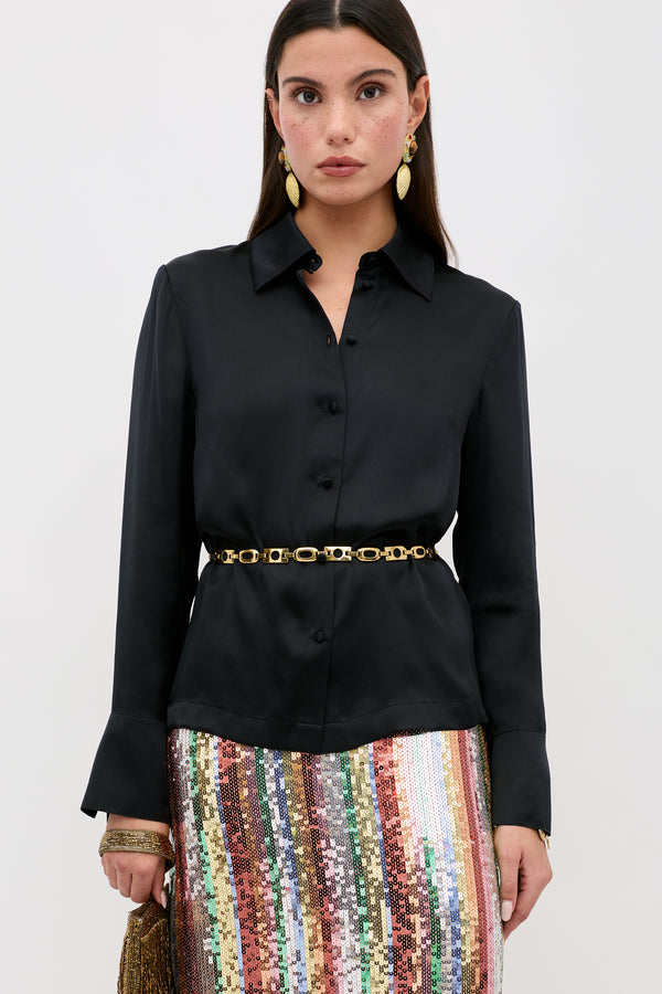 A woman wears the Havan - Black silk button-up shirt with a gold chain belt, a multicolored sequin skirt, gold earrings, and holds a gold clutch against a plain white background.