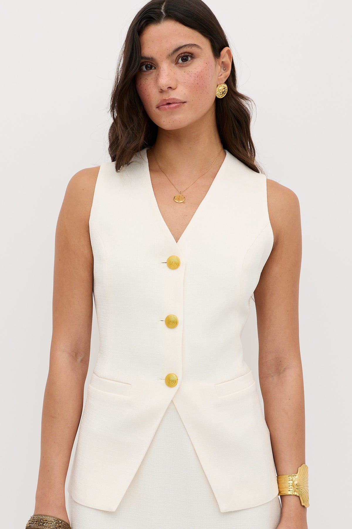 A woman with wavy brown hair wears the Ingrida - Pearl sleeveless white vest with gold buttons and gold jewelry, standing against a plain light background.