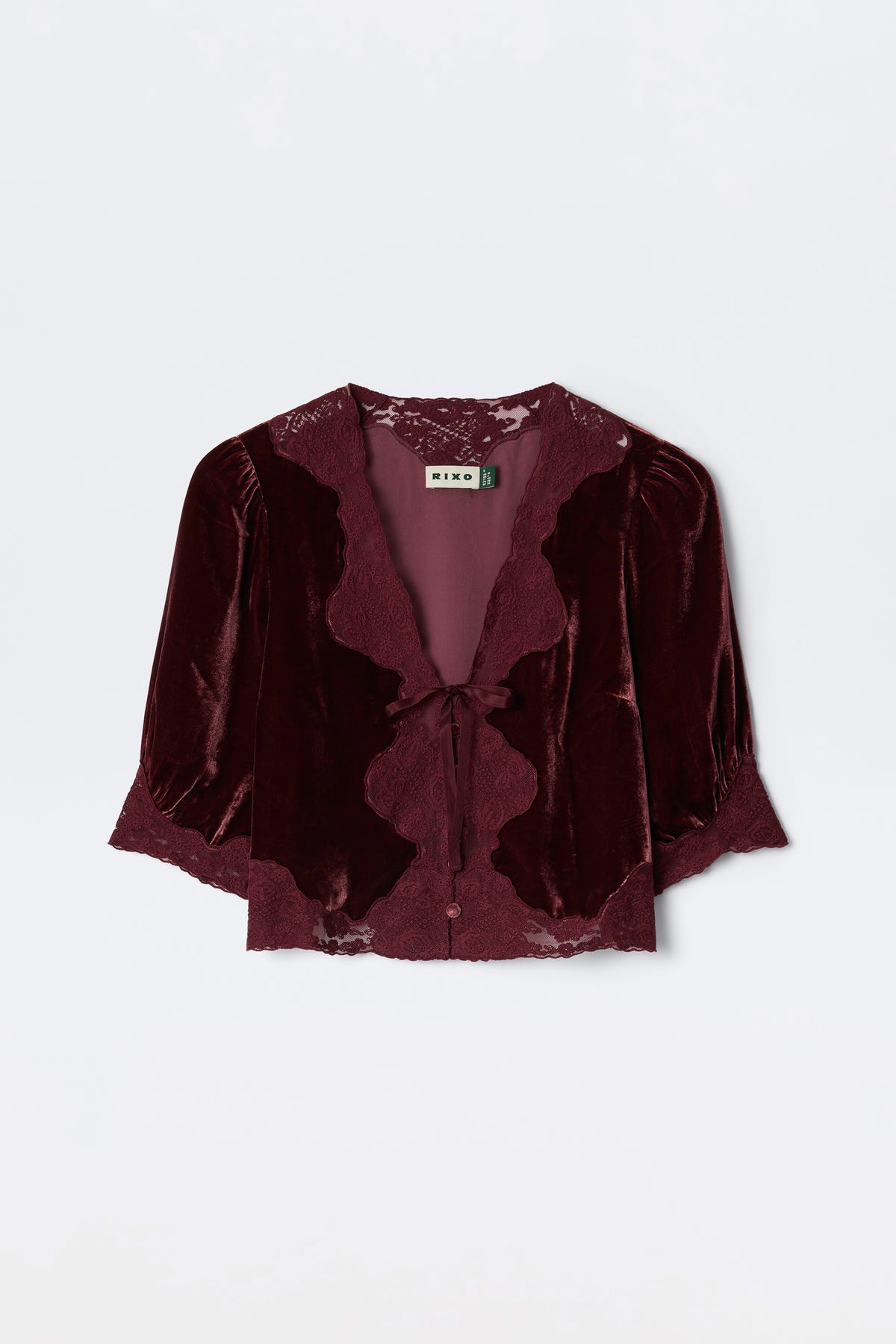 The Ursula - Bordeaux is a burgundy silk-velvet blouse featuring lace trim, puffed mid-length sleeves, and a front tie closure. Vintage-inspired, it’s finished with a “RIXO” label at the neckline.