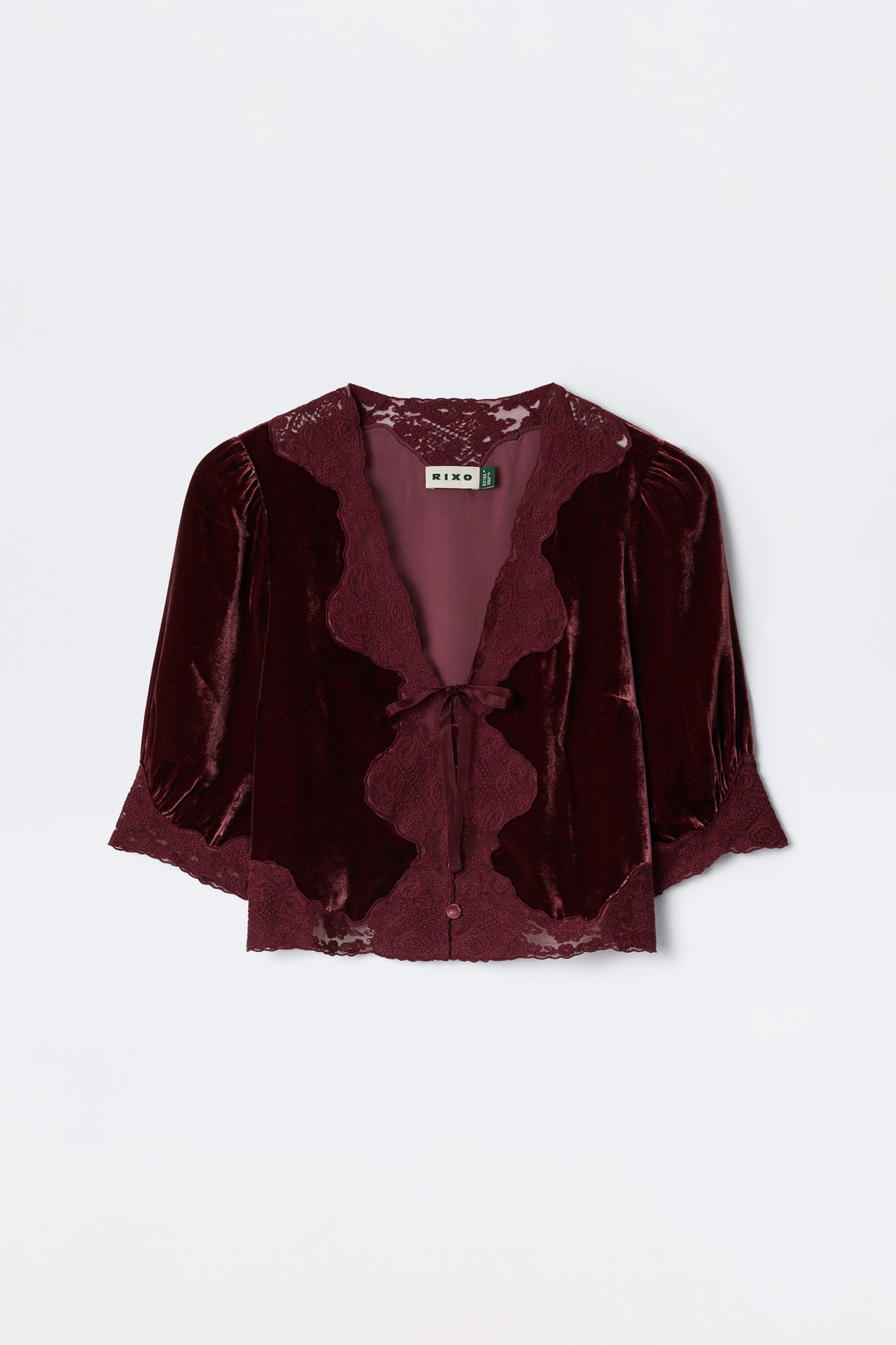 The Ursula - Bordeaux is a burgundy silk-velvet blouse featuring lace trim, puffed mid-length sleeves, and a front tie closure. Vintage-inspired, it’s finished with a “RIXO” label at the neckline.