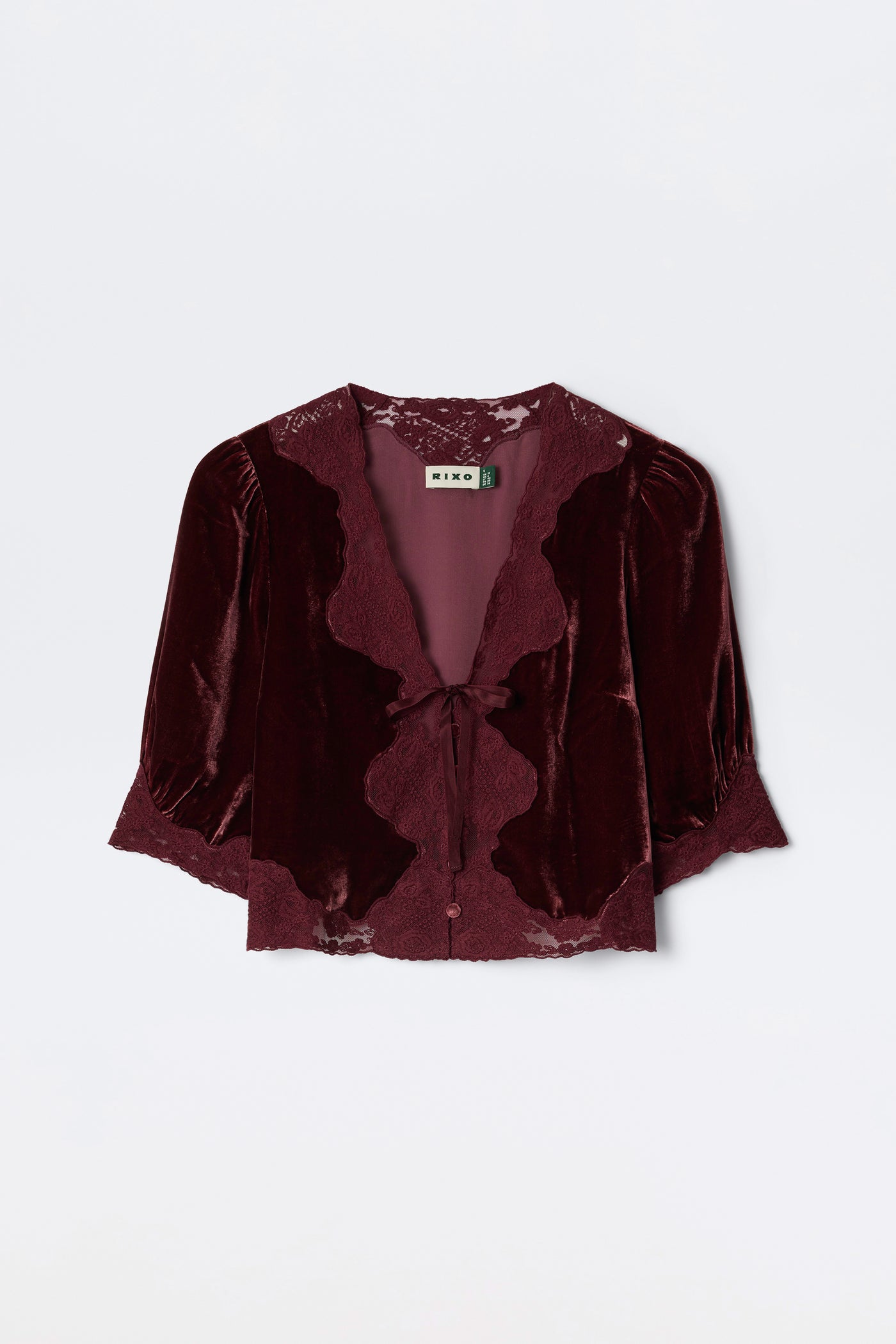 The Ursula - Bordeaux is a burgundy silk-velvet blouse featuring lace trim, puffed mid-length sleeves, and a front tie closure. Vintage-inspired, it’s finished with a “RIXO” label at the neckline.