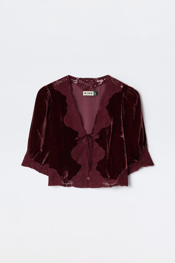 The Ursula - Bordeaux is a burgundy silk-velvet blouse featuring lace trim, puffed mid-length sleeves, and a front tie closure. Vintage-inspired, it’s finished with a “RIXO” label at the neckline.