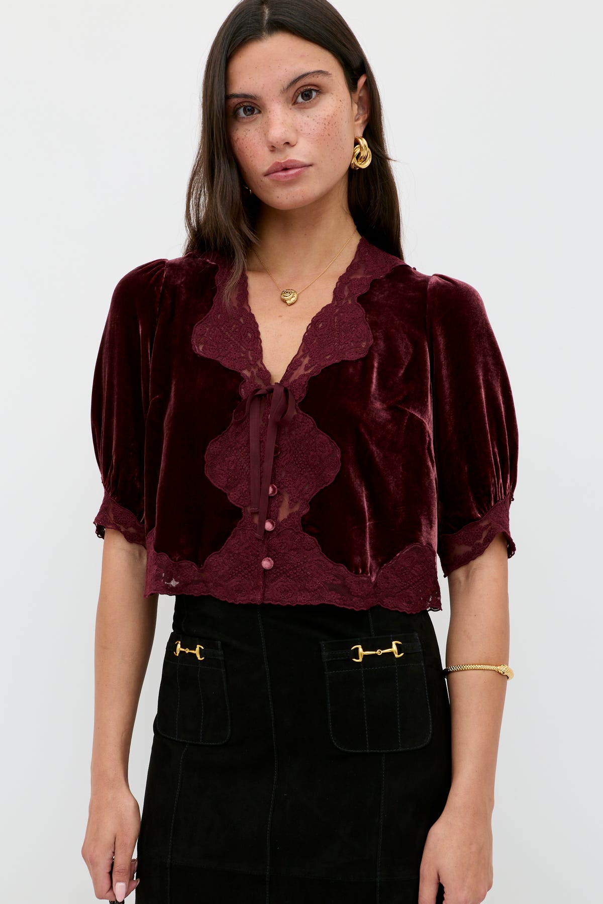 A woman with long dark hair and gold jewelry wears the Ursula - Bordeaux vintage-inspired silk-velvet blouse with lace trim and neck bow, paired with a black skirt featuring gold-accented front pockets, standing against a light background.