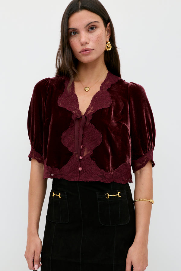 A woman with long dark hair and gold jewelry wears the Ursula - Bordeaux vintage-inspired silk-velvet blouse with lace trim and neck bow, paired with a black skirt featuring gold-accented front pockets, standing against a light background.