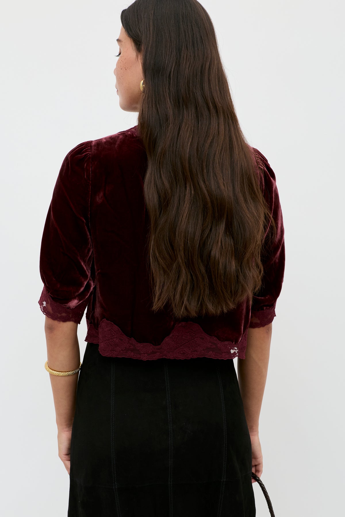 A woman with long, wavy brown hair faces away, wearing the Ursula - Bordeaux vintage-inspired velvet top with scalloped edges and a black skirt. She accessorizes with a gold bracelet and hoop earrings against a plain white background.