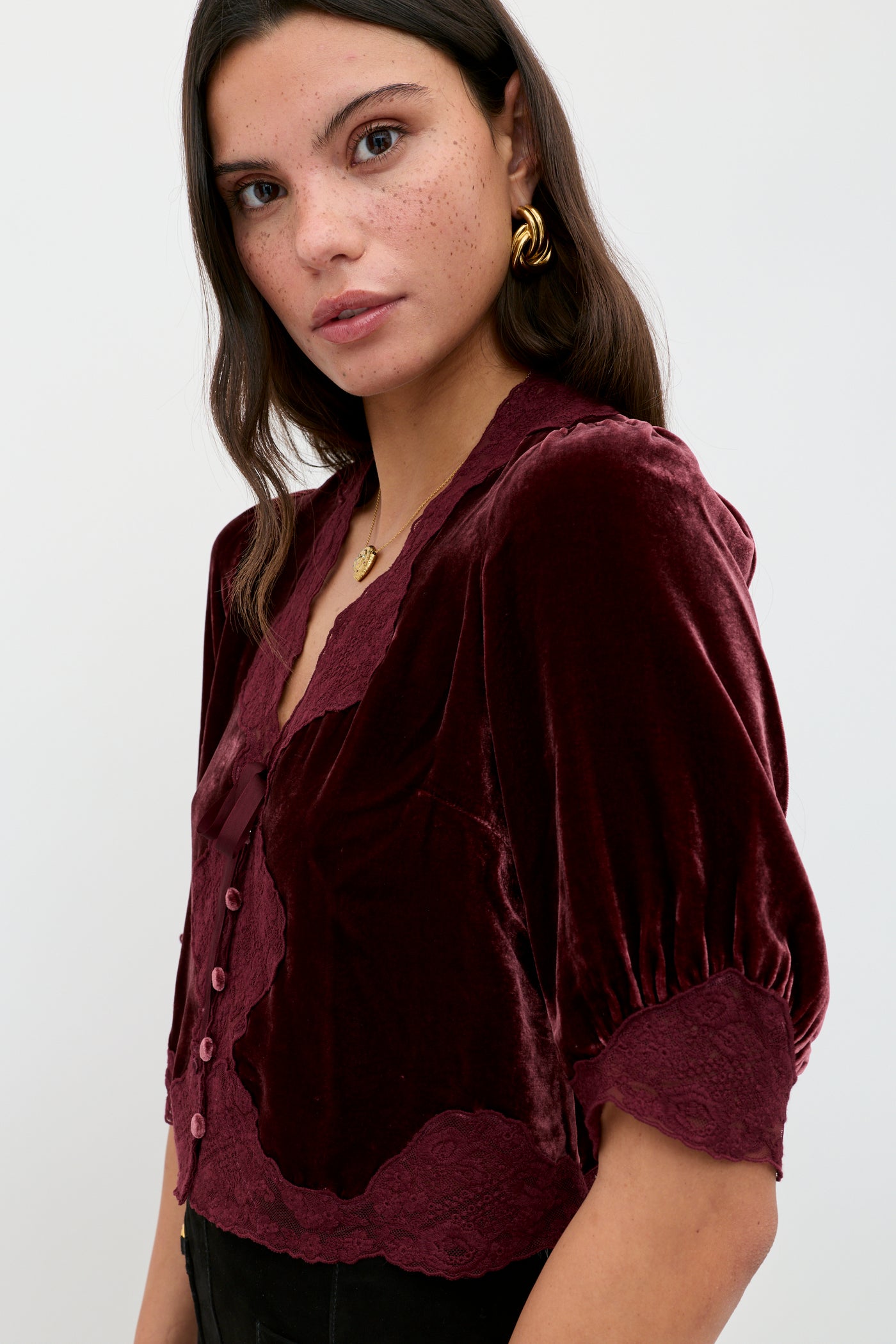 A woman with long dark hair, gold hoop earrings, and a gold necklace wears the Ursula - Bordeaux silk-velvet blouse with lace trim and buttons, standing against a plain light background.