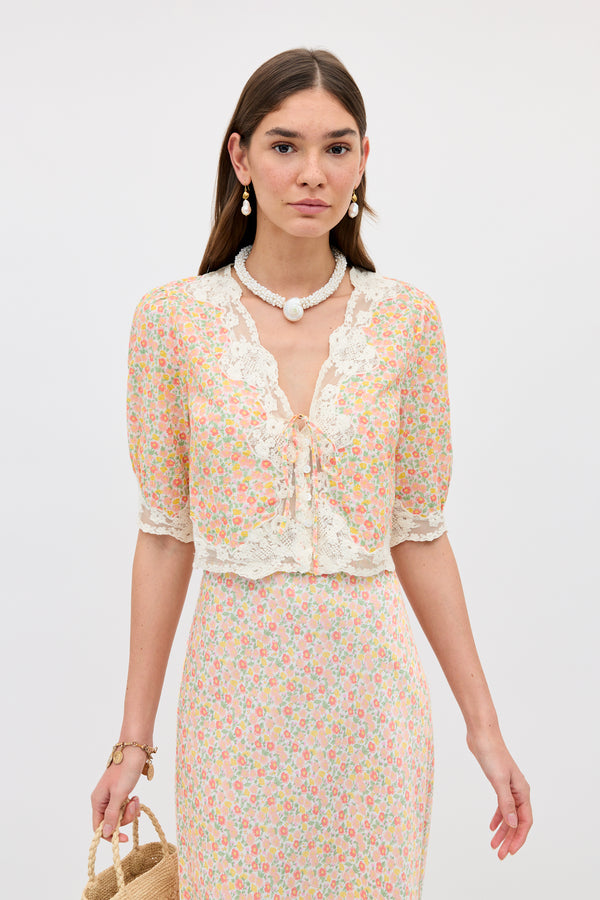 A woman wears the Ursula - Katie Floral Cream cotton top and matching skirt, styled with a straw bag and beaded jewelry, against a simple light background.