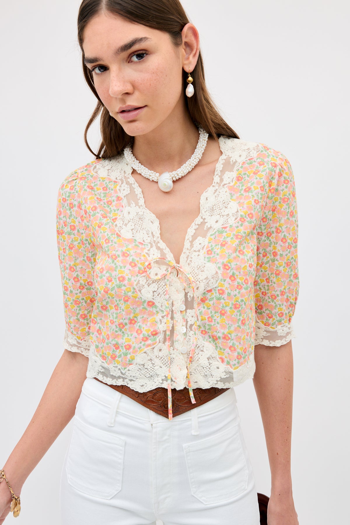 A woman stands against a plain light background wearing the Ursula - Katie Floral Cream vintage-inspired lace top, paired with white high-waisted pants, and accessorized with a beaded pearl necklace and matching earrings.