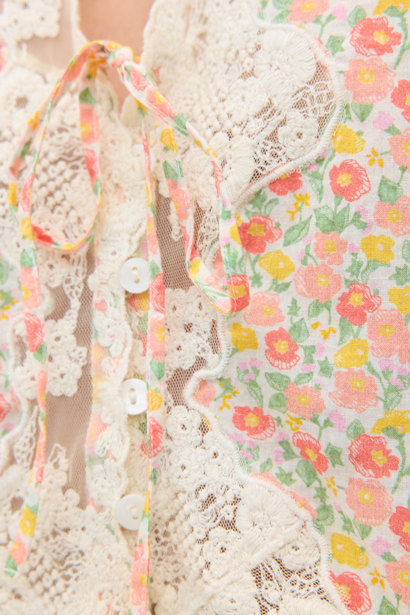 Close-up of the Ursula - Katie Floral Cream top, showcasing its vintage-inspired pink, yellow, and orange floral print on cotton, white lace trim, small buttons, and a fabric tie by the collar with clearly visible textures and details.