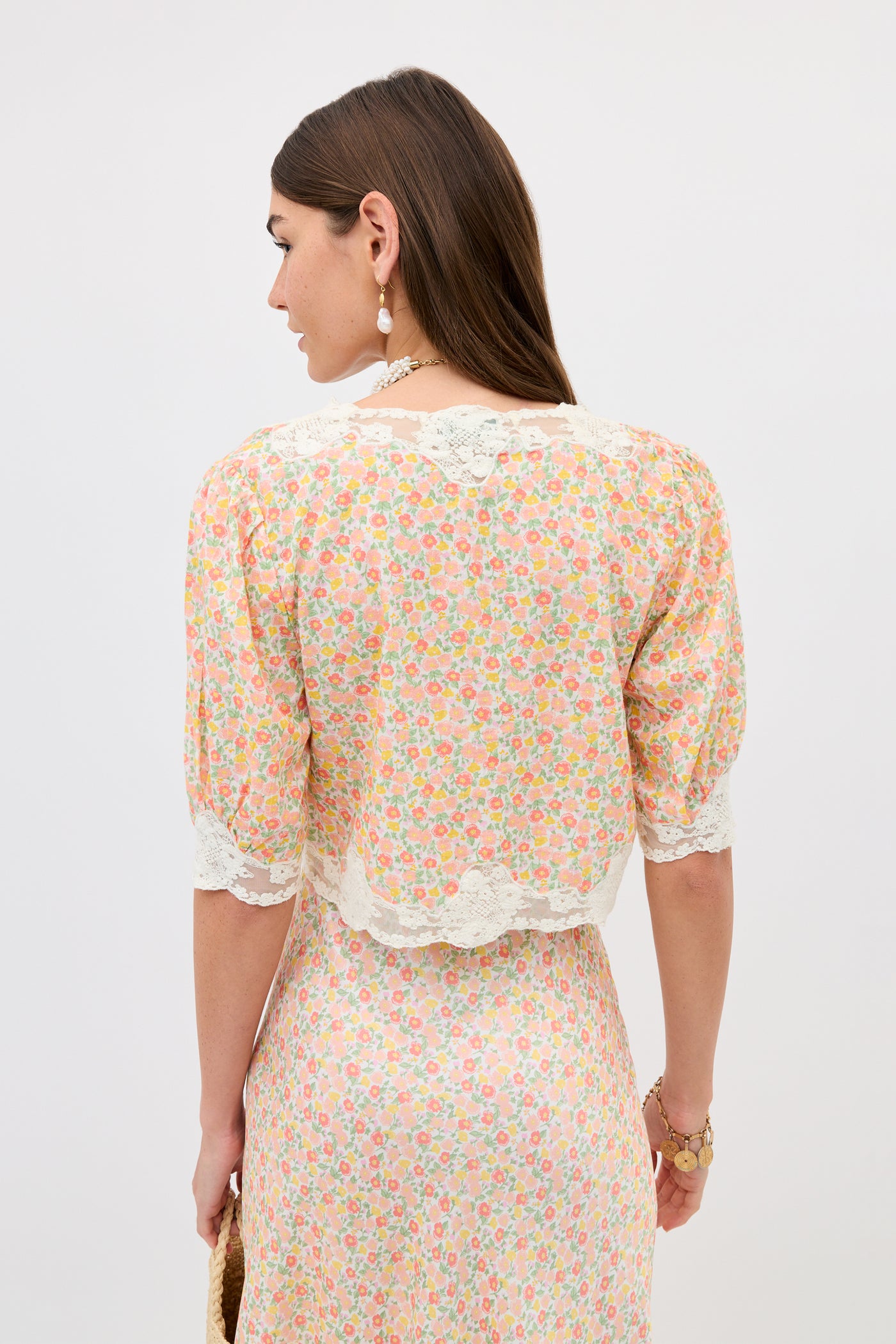 A woman with long brown hair holds a woven bag and wears the Ursula - Katie Floral Cream top, featuring a vintage-inspired floral print, lace trim, and elbow-length sleeves, set against a plain light background.