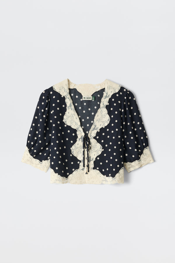 The Ursula - Layla Dot Navy is a navy blouse with white polka dots, puffed half sleeves, cream lace trim on the collar, front, and hem, plus a front tie closure.