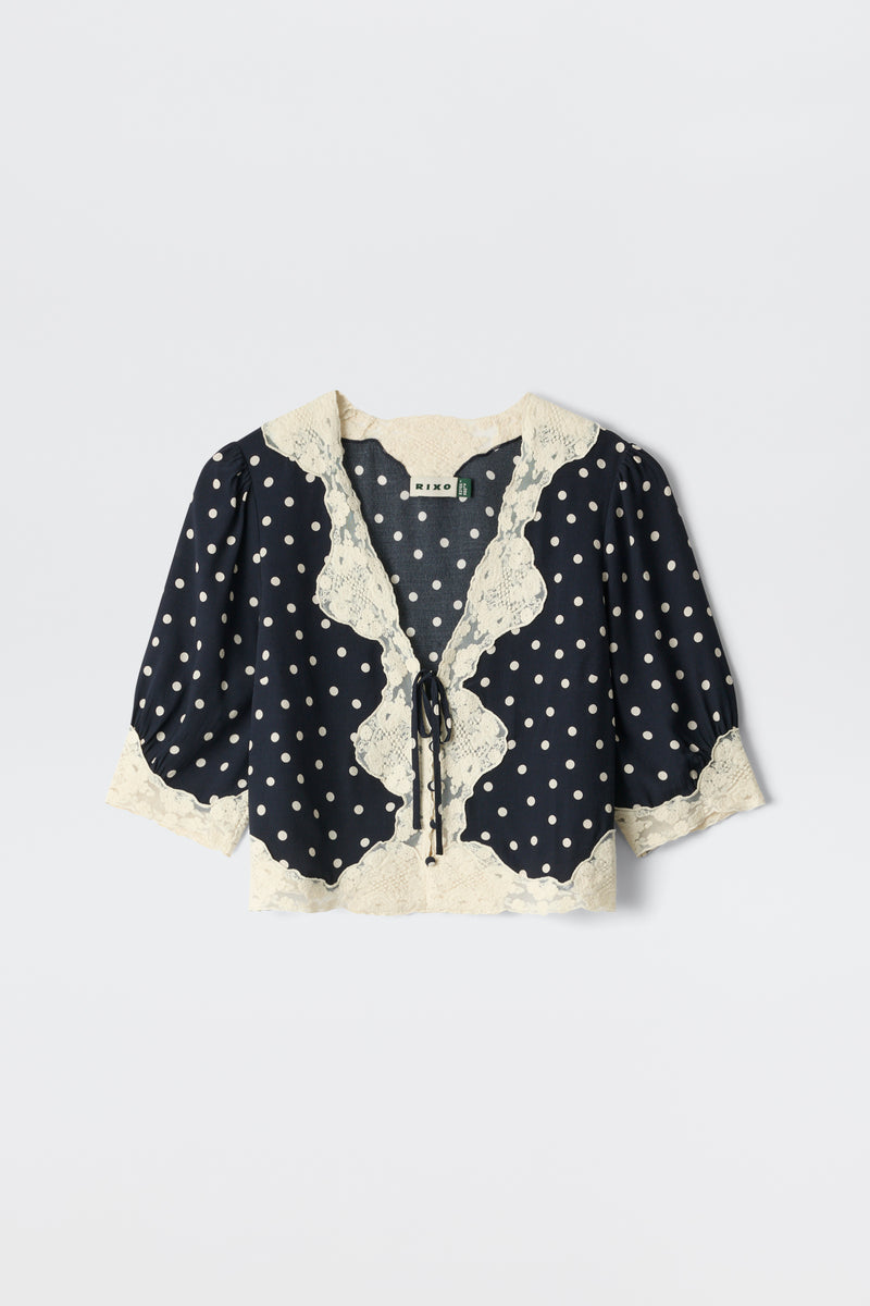 The Ursula - Layla Dot Navy is a navy blouse with white polka dots, puffed half sleeves, cream lace trim on the collar, front, and hem, plus a front tie closure.