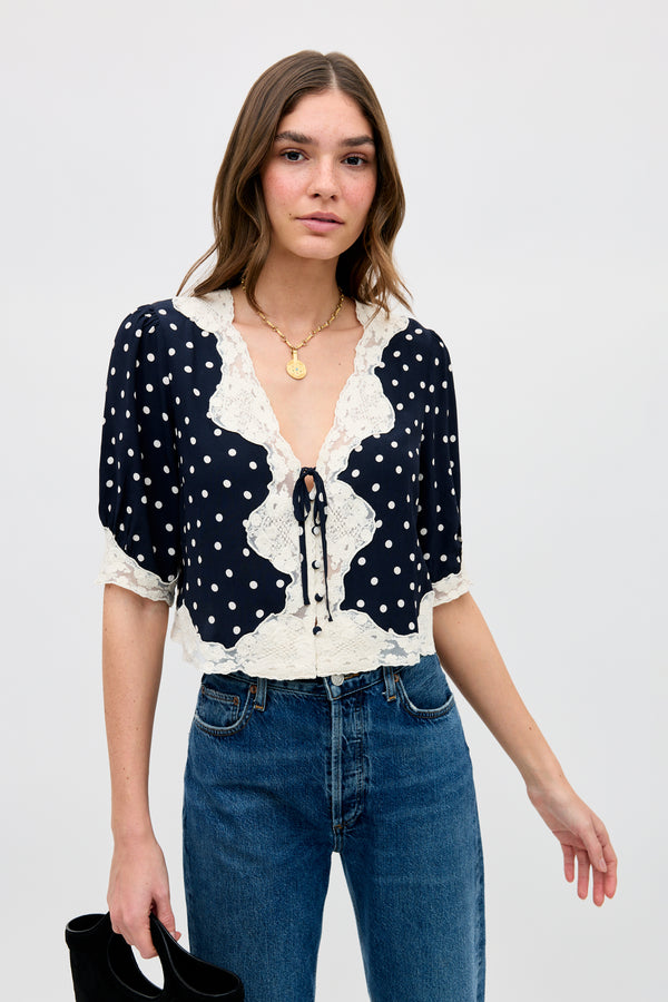 A woman with long brown hair wears the Ursula - Layla Dot Navy blouse, featuring navy polka dots, white lace trim, and puff sleeves. She pairs it with blue jeans and holds a black handbag against a plain light background.