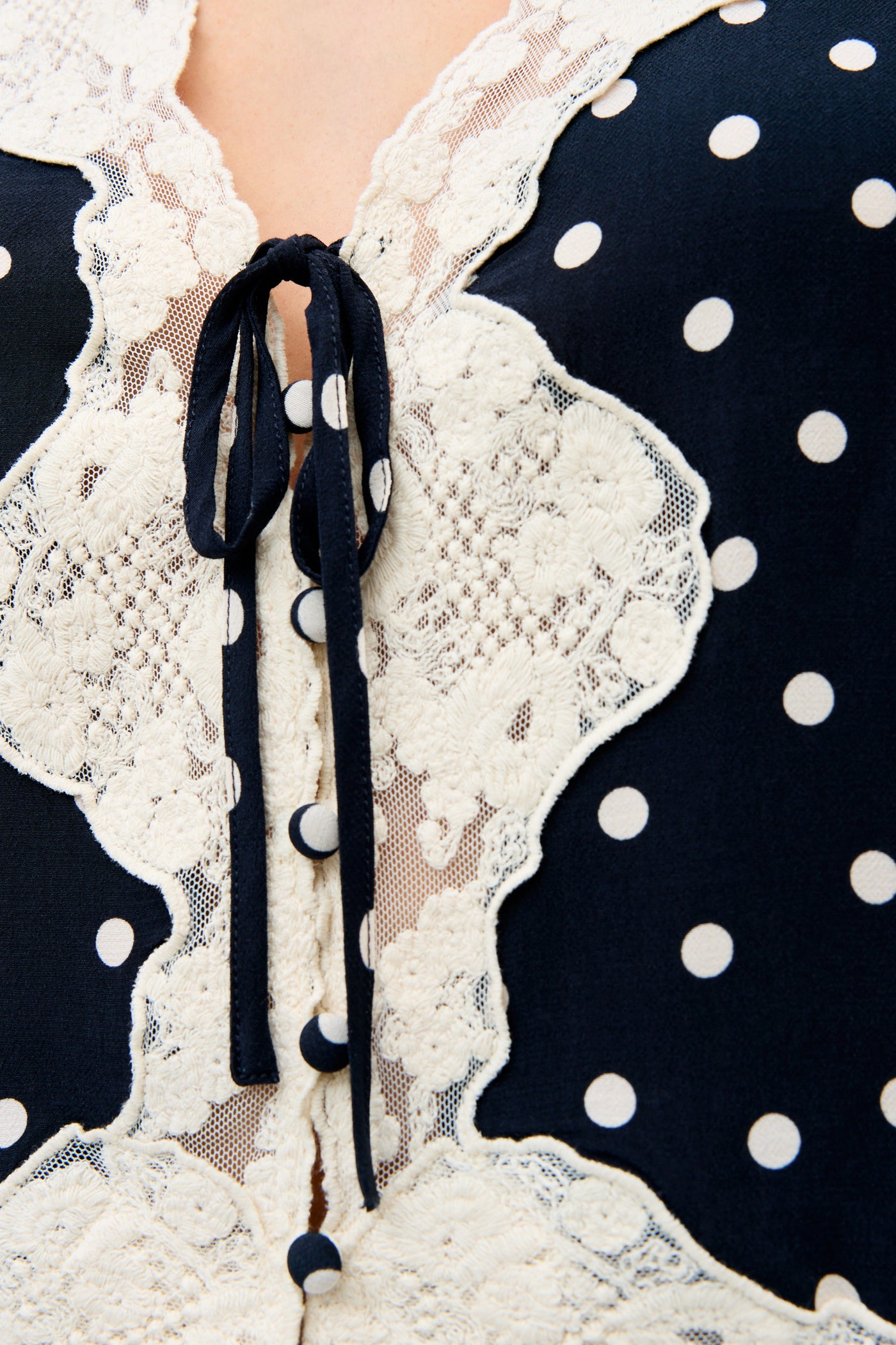 Close-up of the Ursula - Layla Dot Navy, featuring navy blue fabric with white polka dots, intricate white lace trim, fabric-covered buttons, and a tied bow at the neckline for a playful yet elegant look.