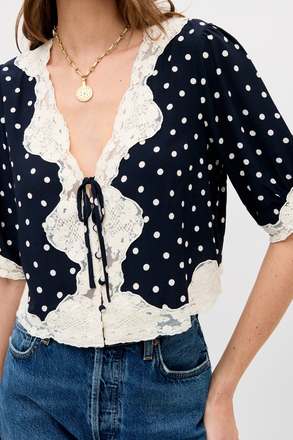 A person wearing the Ursula - Layla Dot Navy blouse—navy blue with short sleeves, polka dots, cream lace trim, and a tie front—paired with blue jeans and a gold chain necklace with a round pendant.