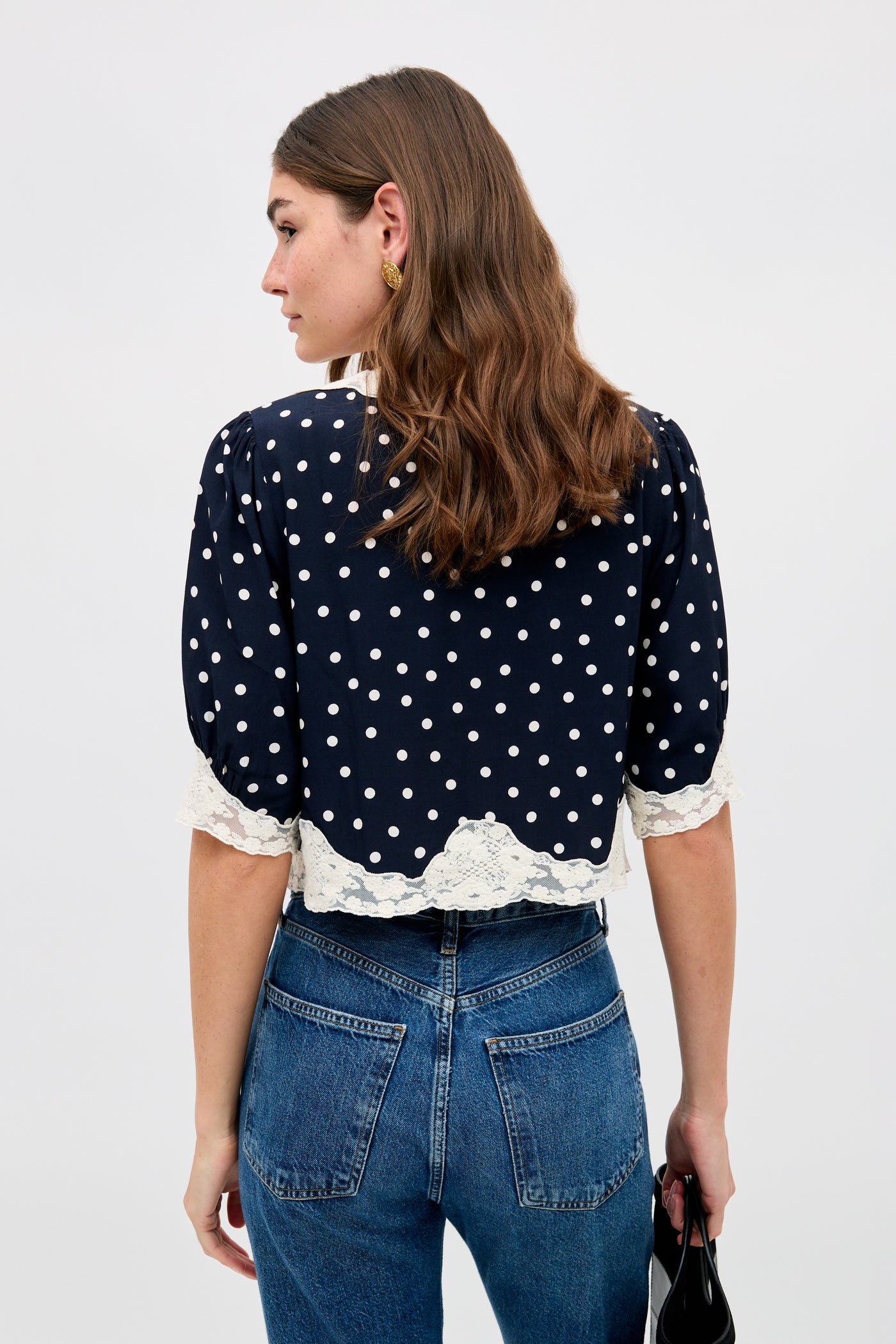 A woman with wavy brown hair is seen from behind wearing the Ursula - Layla Dot Navy blouse with white polka dots, lace trim on the sleeves and hem, paired with high-waisted blue jeans. She holds a black item in her right hand.