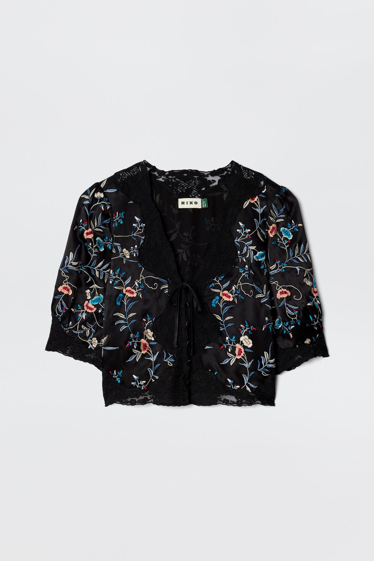 The Ursula - Botanical Embroidery Black is a silky satin short-sleeve blouse with lace trim, tie-front closure, and colorful botanical embroidery. It features a RIXO brand label inside the collar.
