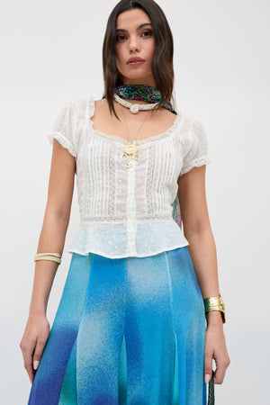 A woman wears the Sandi - Ivory blouse, featuring short sleeves, broderie anglaise cotton with lace inserts, and a square neckline, styled with a blue gradient skirt and green scarf—ideal for everyday wear against a plain background.