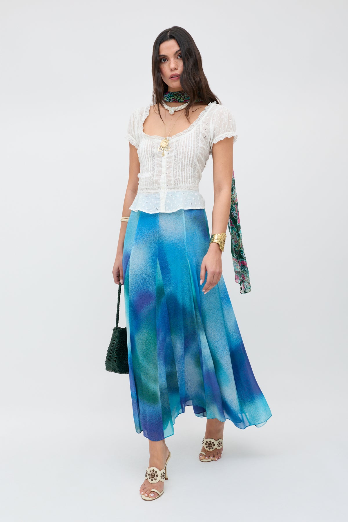 A woman poses against a plain background, wearing the Sandi - Ivory white broderie anglaise cotton blouse with lace inserts, paired with a blue and green flowy skirt, floral sandals, gold jewelry, a green woven bag, and a green floral neck scarf.