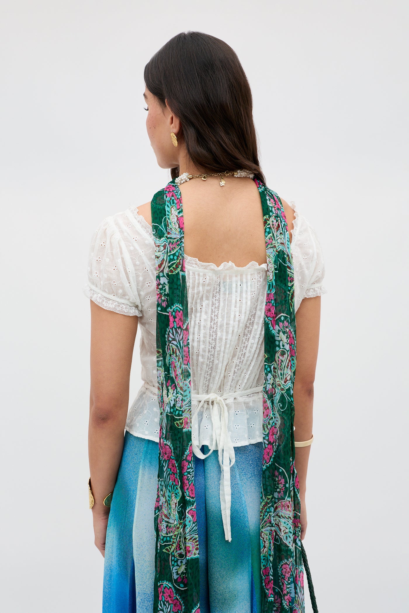 A woman faces away, wearing the Sandi - Ivory broderie anglaise cotton blouse with puff sleeves, a blue-gradient skirt, green floral scarf, gold jewelry, and flowing dark hair—an effortlessly chic everyday look.