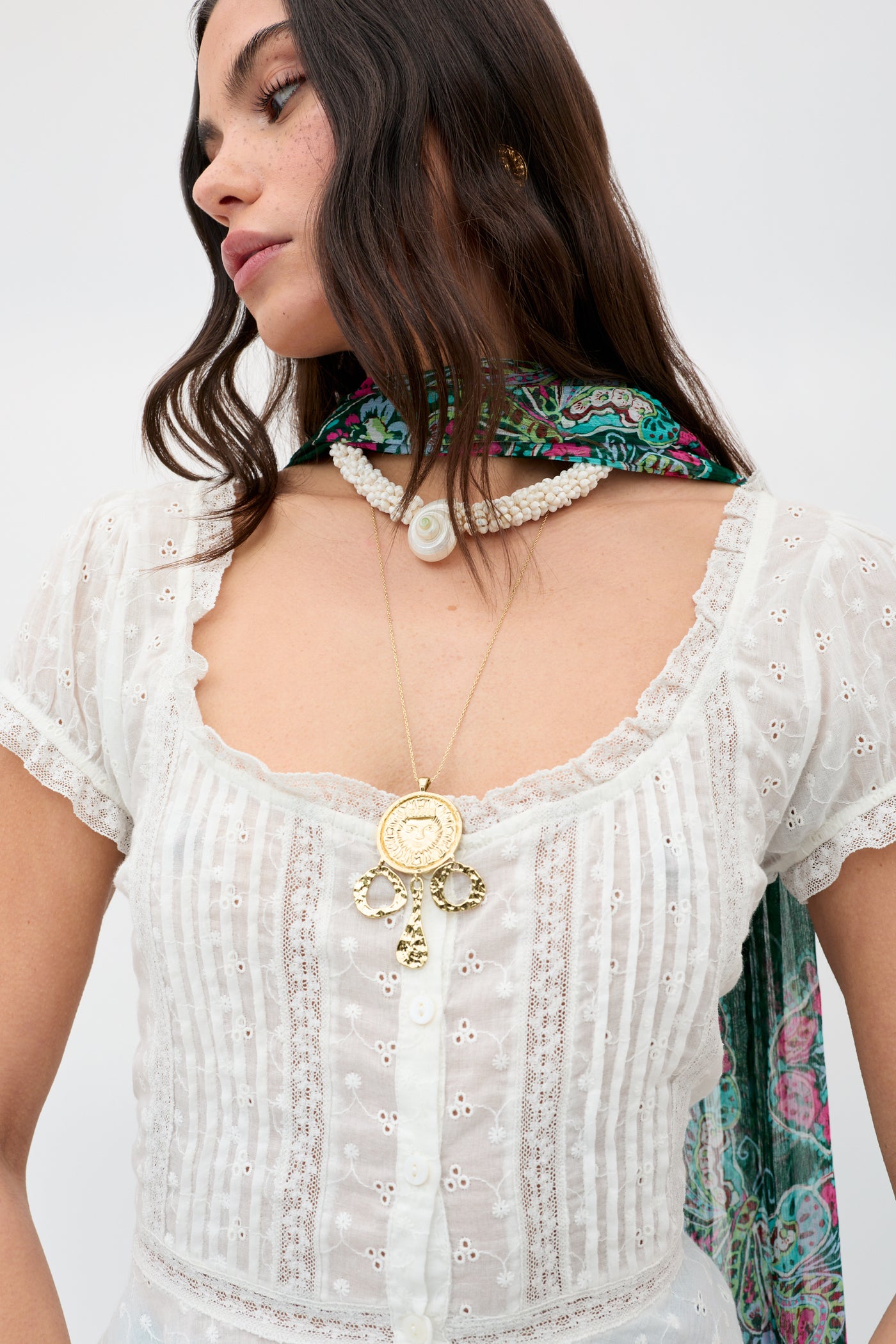A woman wears the Sandi - Ivory blouse, a white broderie anglaise cotton top with vertical stripes and a square neckline. She styles it with layered necklaces, a floral scarf over her shoulder, and loose brown hair for effortless style.