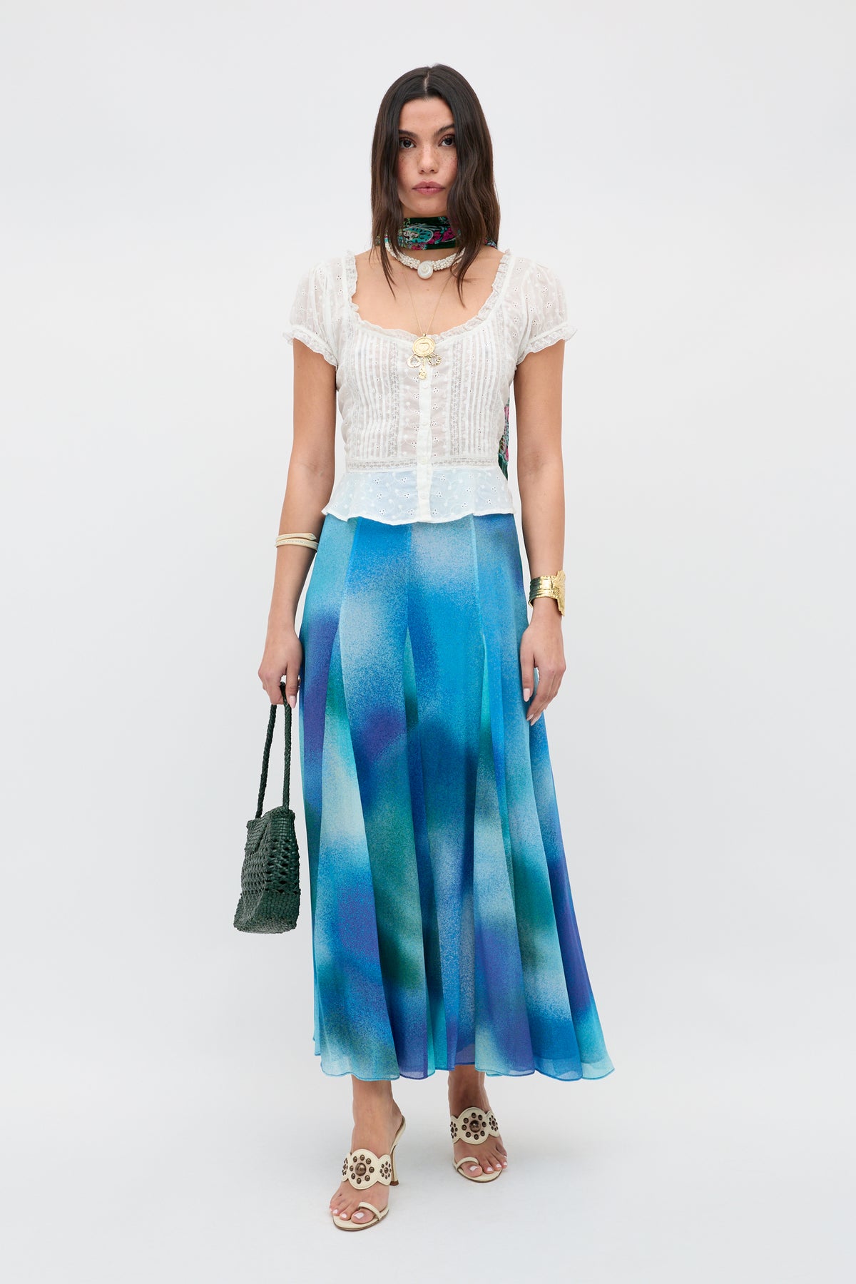 A woman poses against a white background wearing the Sandi - Ivory broderie anglaise cotton blouse with a blue gradient maxi skirt, green scarf, white sandals, gold jewelry, and carrying a green woven bag for effortless style.