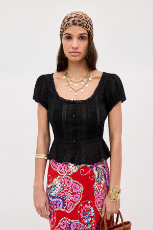 A woman in the Sandi - Black broderie anglaise cotton short-sleeve blouse, a leopard print headscarf, gold layered necklaces, and a red paisley skirt stands against a plain background.