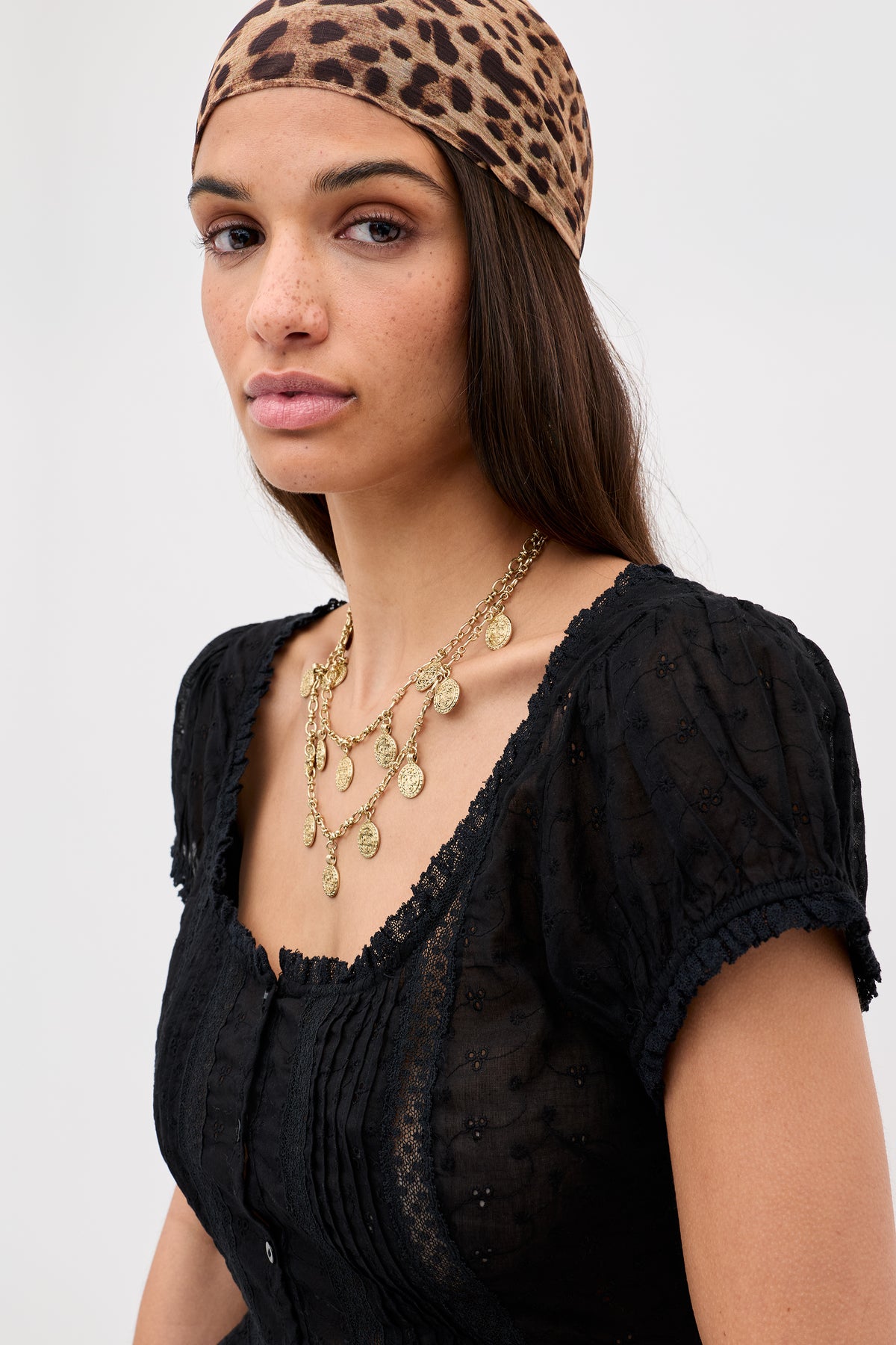 A woman with long dark hair wears the Sandi - Black blouse featuring broderie anglaise cotton and lace details, paired with a leopard print headscarf and gold coin necklaces, standing against a plain light background.