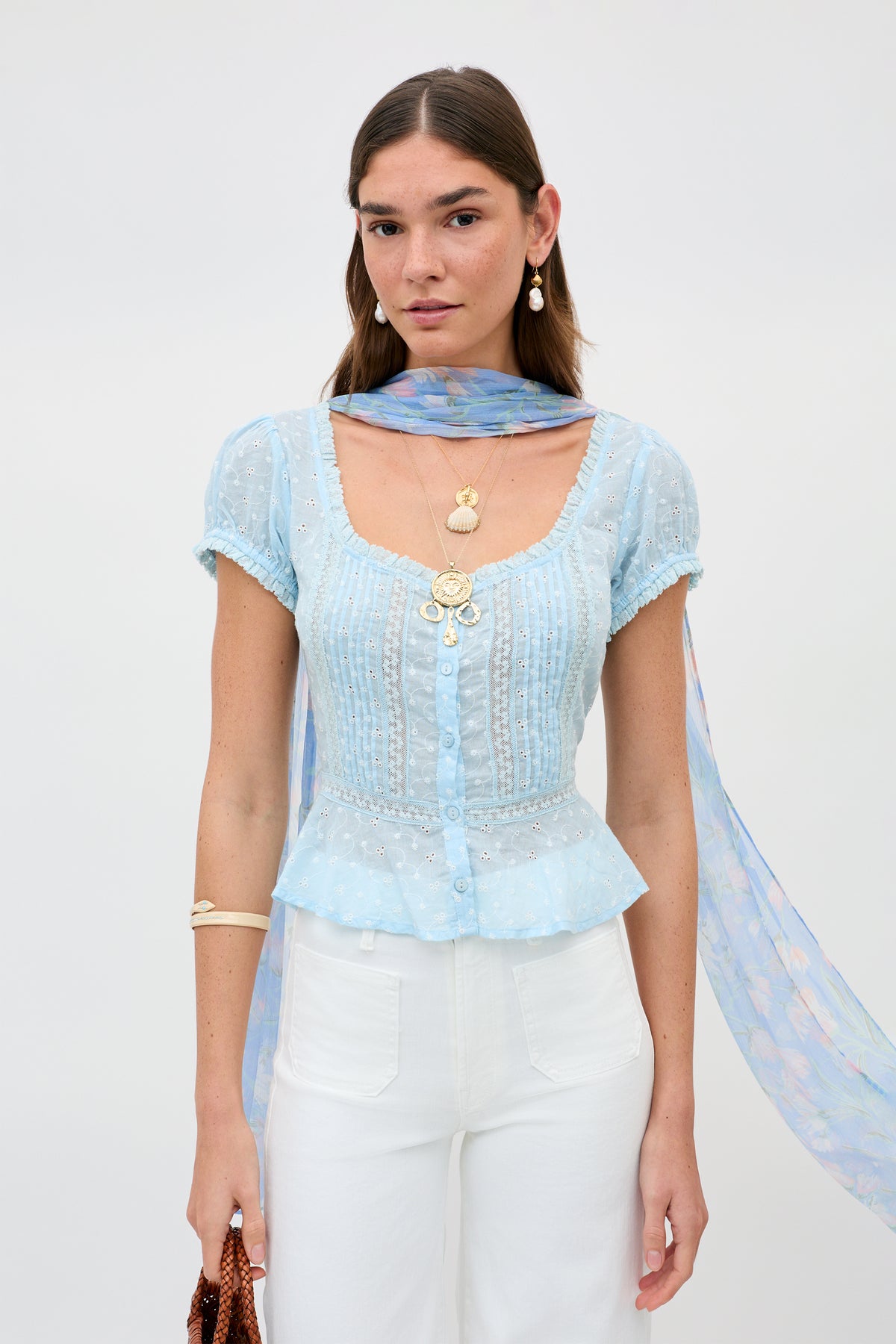 A woman wears the Sandi - Powder Blue top with delicate white patterns, paired with white high-waisted pants, layered gold necklaces, earrings, a sheer blue scarf around her neck, and holds a brown textured bag.