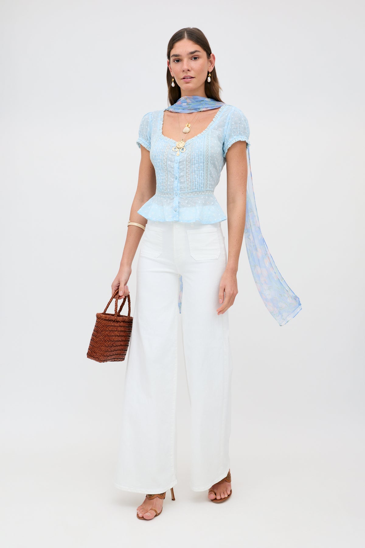 A woman stands against a white background wearing Sandi - Powder Blue, paired with white wide-leg pants, a sheer blue scarf, sandals, gold jewelry, and holding a brown woven handbag.