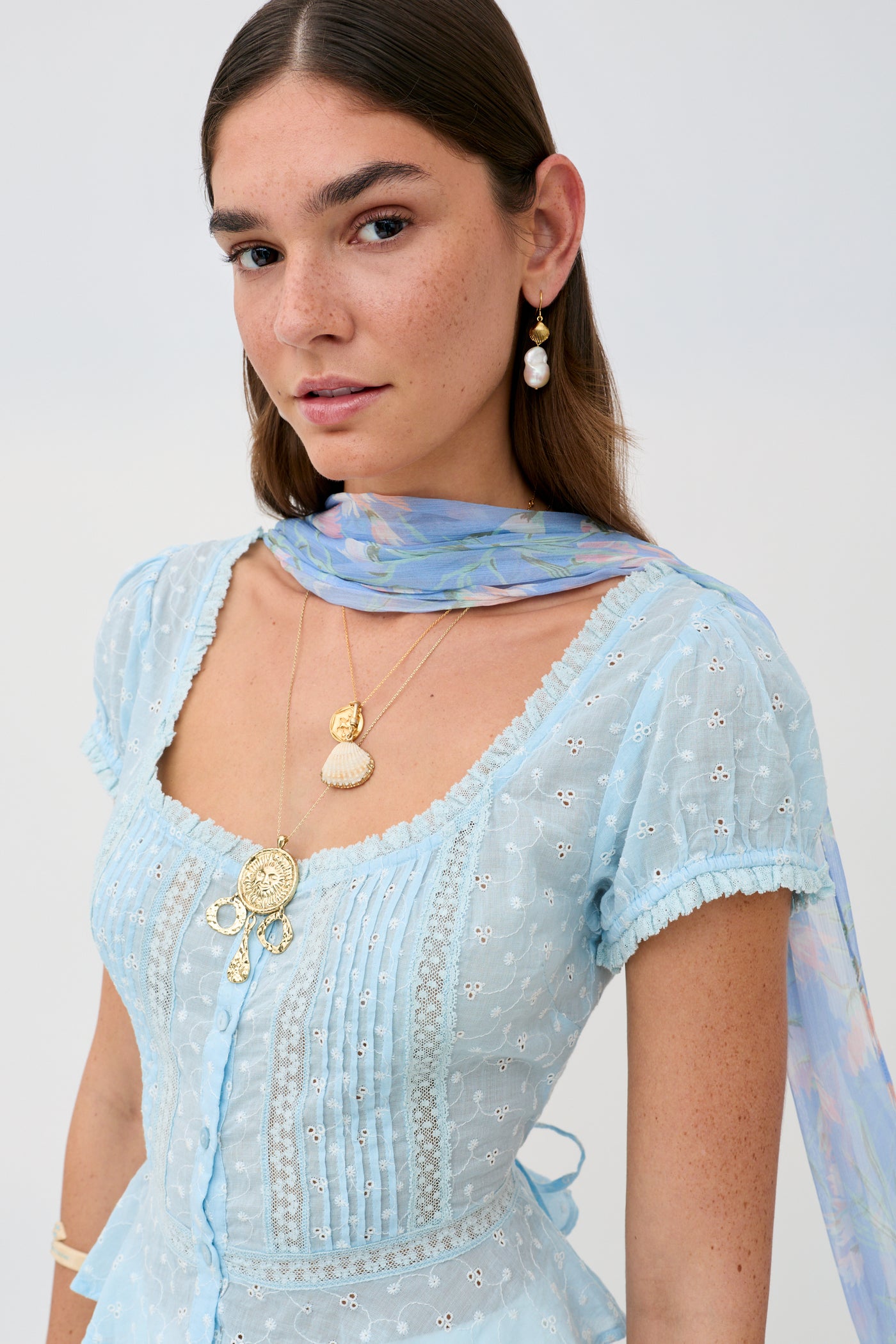 A woman with long brown hair wears Sandi - Powder Blue, styled with layered gold necklaces and pearl earrings, and poses with a sheer pastel scarf draped over her shoulders against a light background.