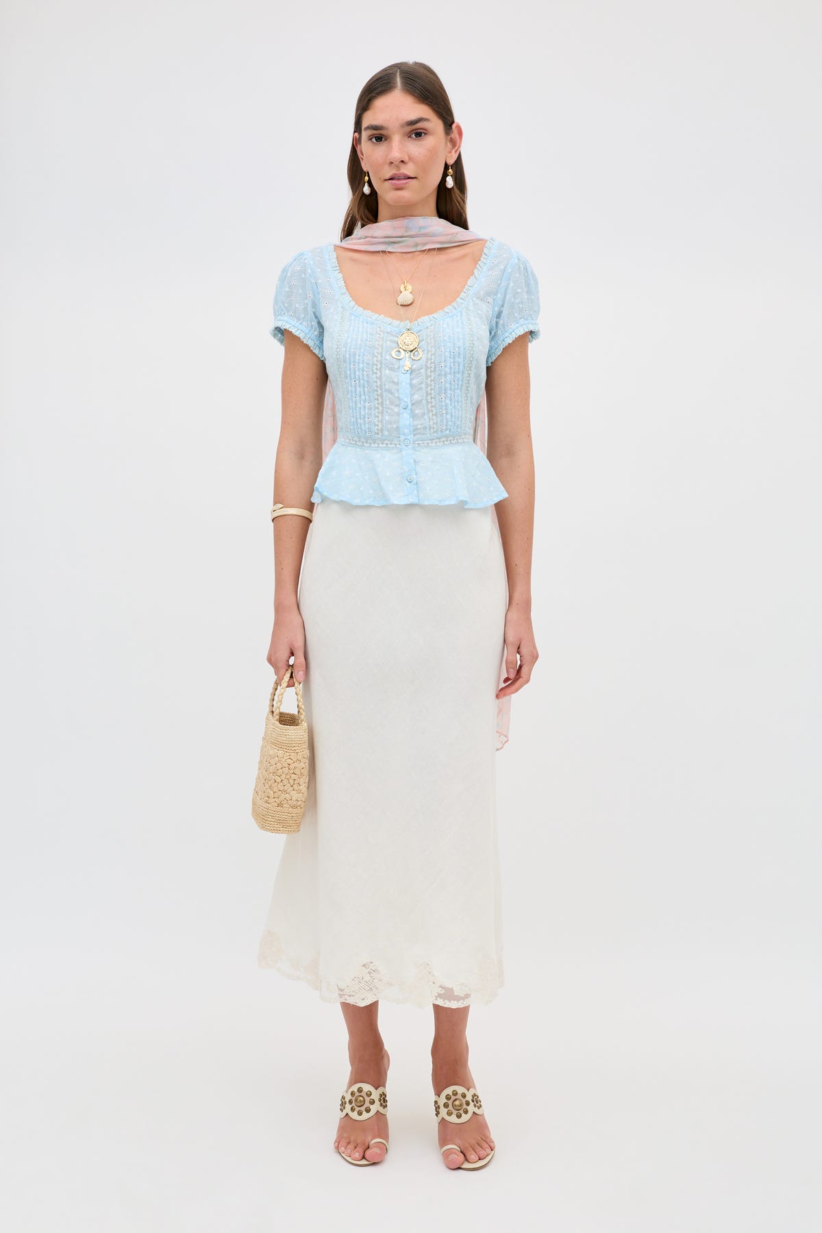 A woman stands against a plain background wearing Sandi - Powder Blue, a peplum hem top paired with a white lace-insert midi skirt, tan sandals, layered necklaces, and holding a woven handbag.