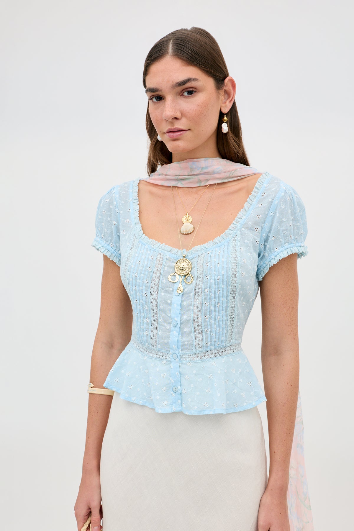 A woman with straight brown hair wears the Sandi - Powder Blue broderie anglaise cotton top with lace inserts, gold layered necklaces, earrings, a pink scarf, and a cream skirt against a plain background.