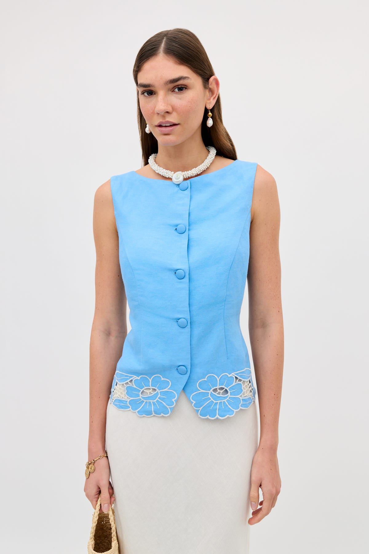 A woman stands against a plain light background, holding a woven bag and wearing the Lucca - Daisy Cutwork Embroidery Blue sleeveless linen top with cutwork floral hem, styled with a pearl necklace, drop earrings, and a white skirt.