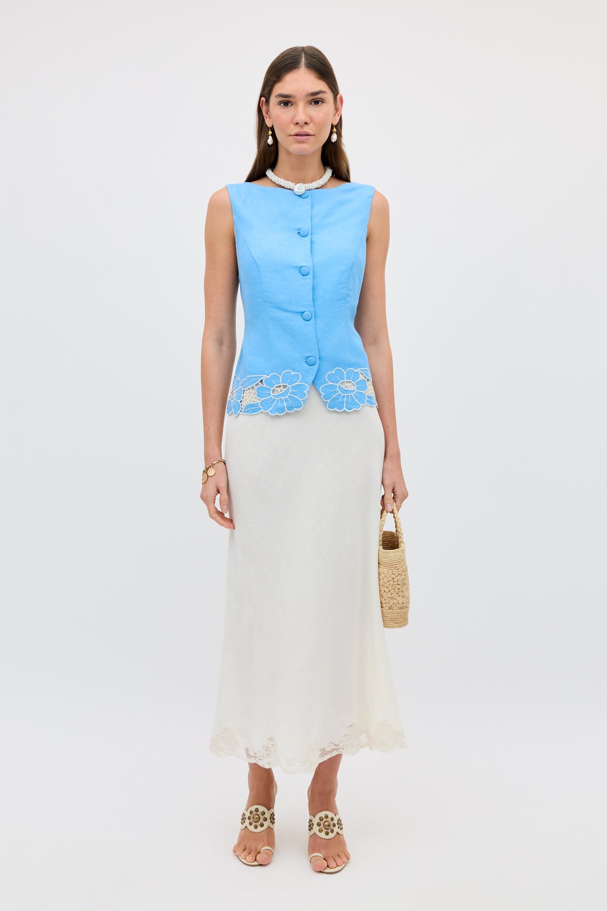 A woman stands against a plain background, wearing the Lucca - Daisy Cutwork Embroidery Blue linen top, a long white skirt with lace hem, beige heeled sandals, pearl jewelry, and carrying a woven handbag.