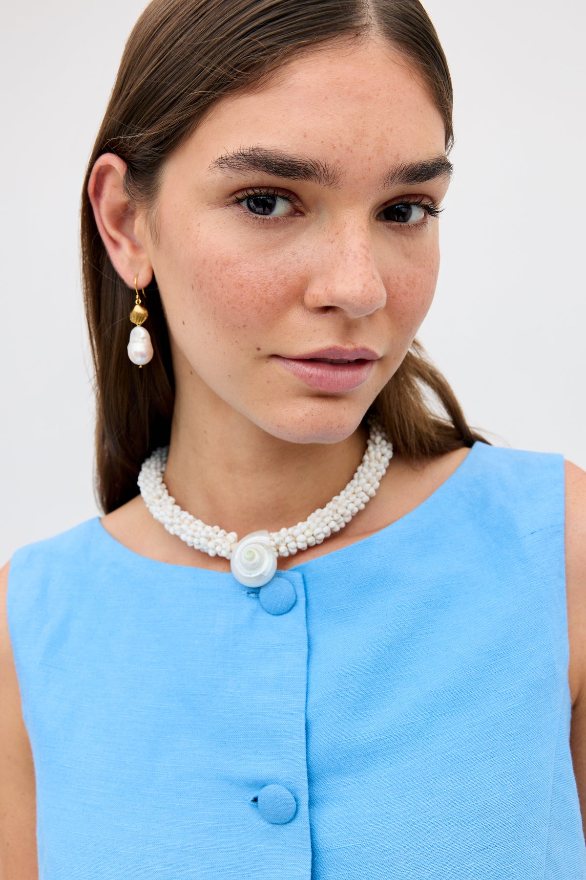 A woman with straight brown hair wears the Lucca - Daisy Cutwork Embroidery Blue sleeveless linen top, a pearl necklace with a white rose pendant, and a dangling pearl earring. She gazes slightly to the side with a neutral expression.