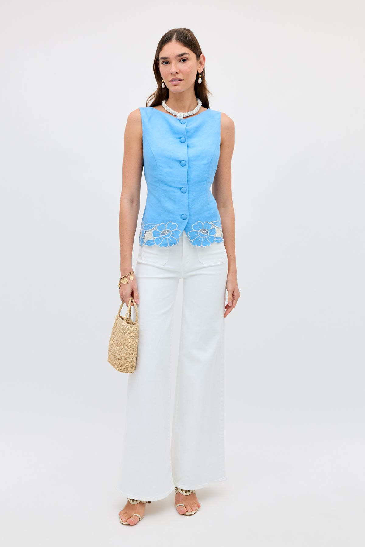 A woman wears the Lucca - Daisy Cutwork Embroidery Blue sleeveless linen top, white wide-leg pants, beige sandals, a pearl necklace, and holds a woven straw handbag while standing against a plain white background.