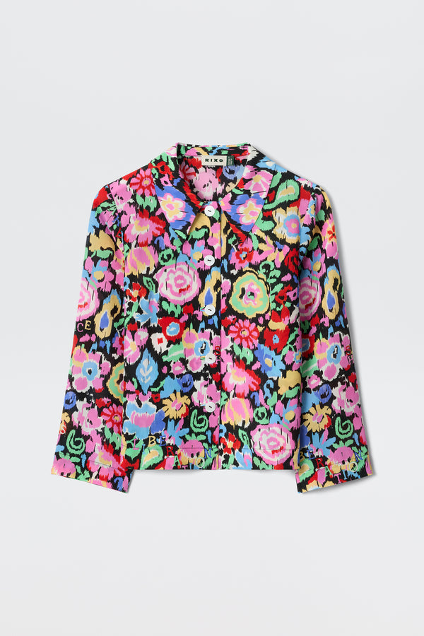 The Ginette - Ikat Bloom Charcoal is a long-sleeve, collared jacket with a vibrant, multicolored floral pattern in pink, blue, green, yellow, and red on a black background, shown against a white surface.
