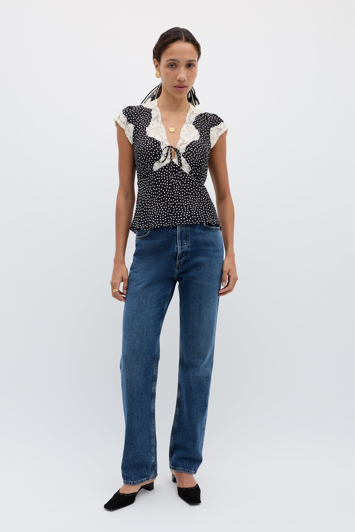 A woman with dark hair pulled back models the Elaine - Mini Polka Dot Black top, featuring black and white polka dots with lace trim. She pairs it with blue high-waisted jeans, black pointed-toe shoes, and gold jewelry against a plain background.
