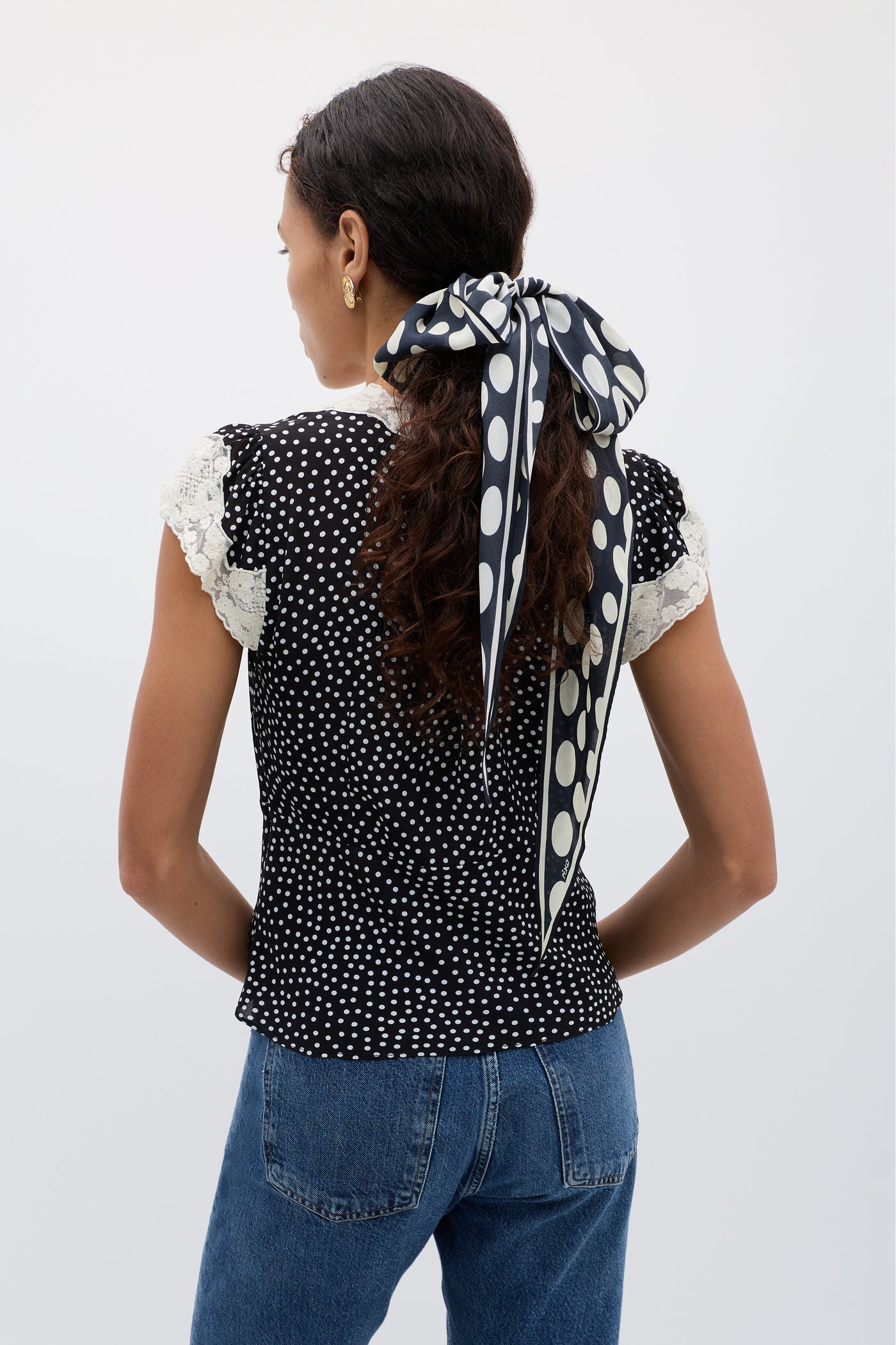 A woman with curly hair, seen from behind in blue jeans and a vintage black blouse, ties her hair with the Elaine - Mini Polka Dot Black scarf.