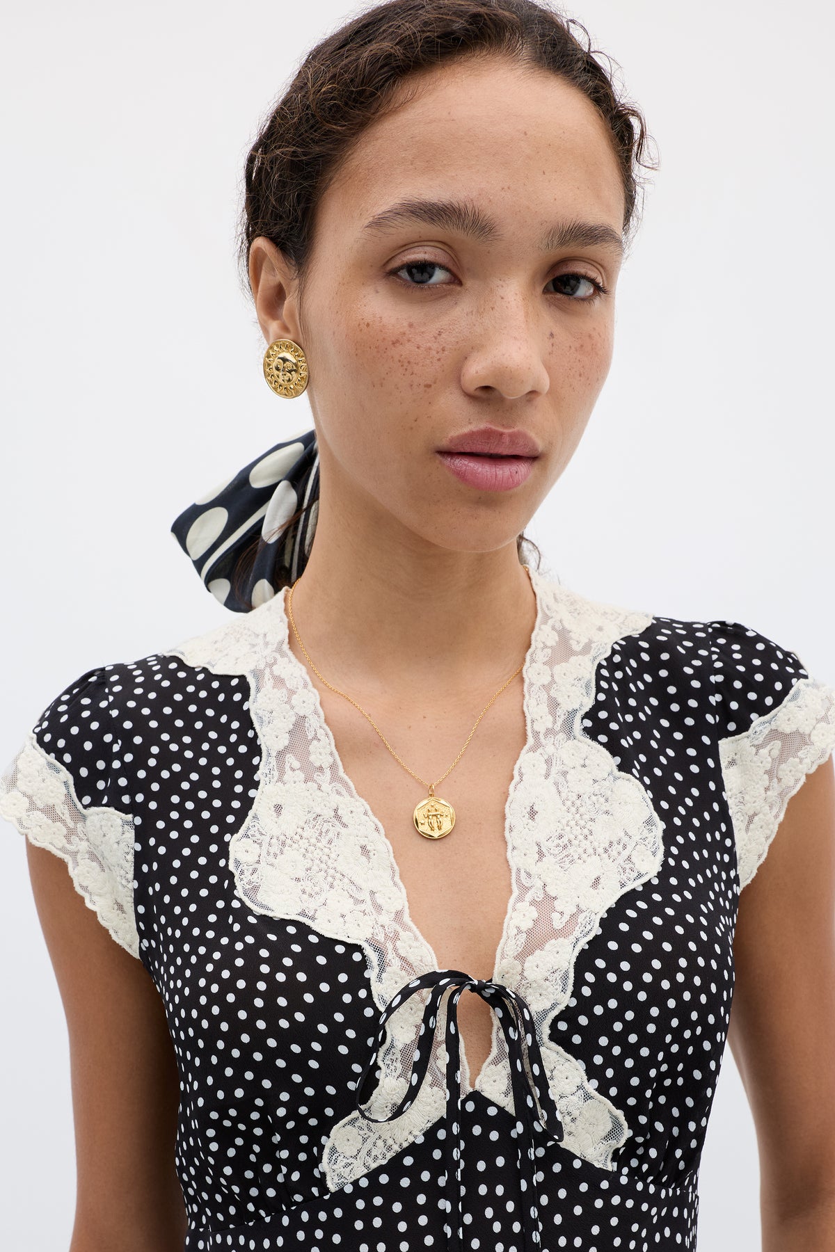 Elaine poses against a light background wearing the "Elaine - Mini Polka Dot Black" dress with lace trim, accessorized with a gold pendant necklace, gold earrings, and a matching polka dot scarf styled in her hair.