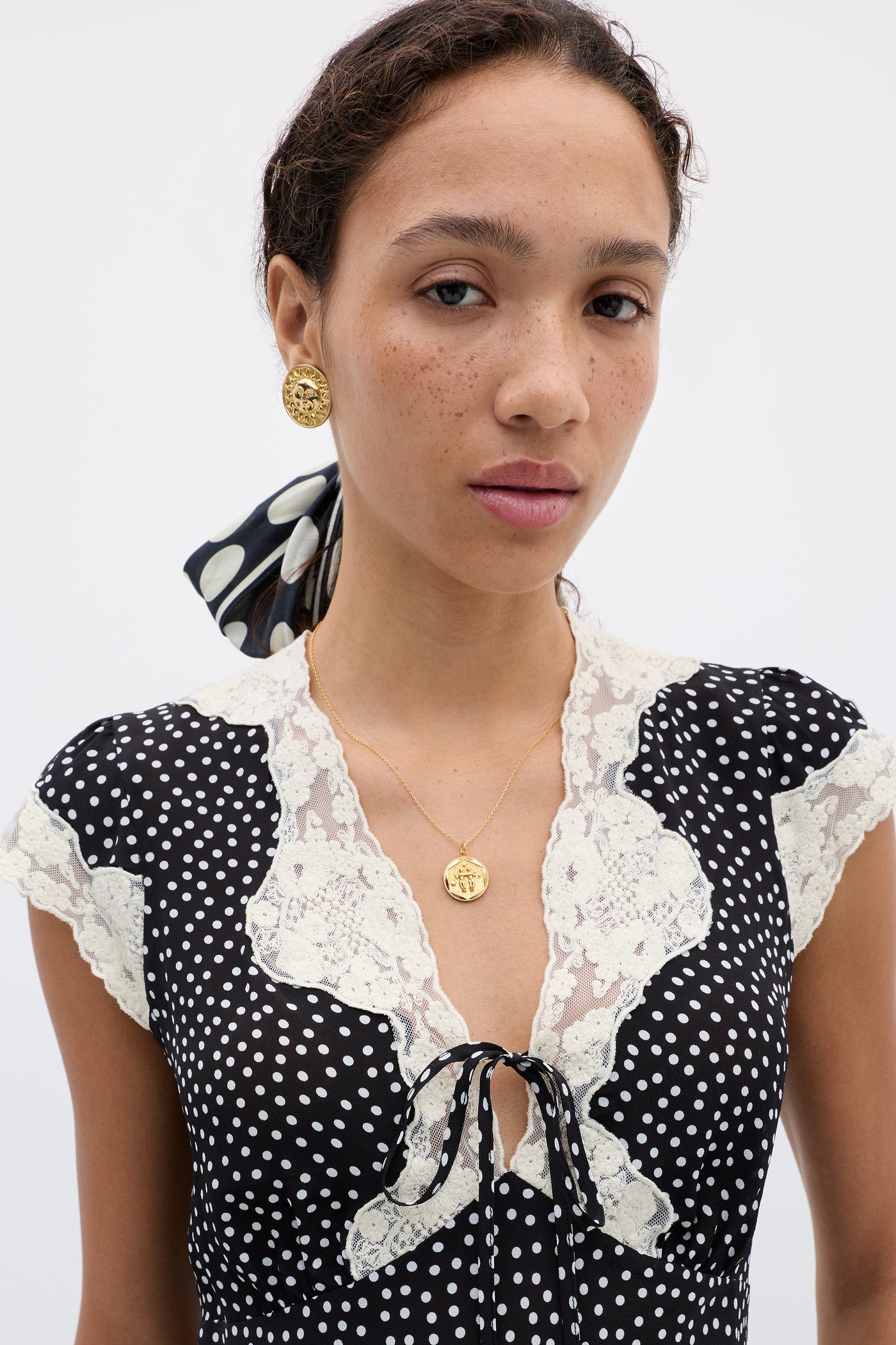 Elaine poses against a light background wearing the "Elaine - Mini Polka Dot Black" dress with lace trim, accessorized with a gold pendant necklace, gold earrings, and a matching polka dot scarf styled in her hair.