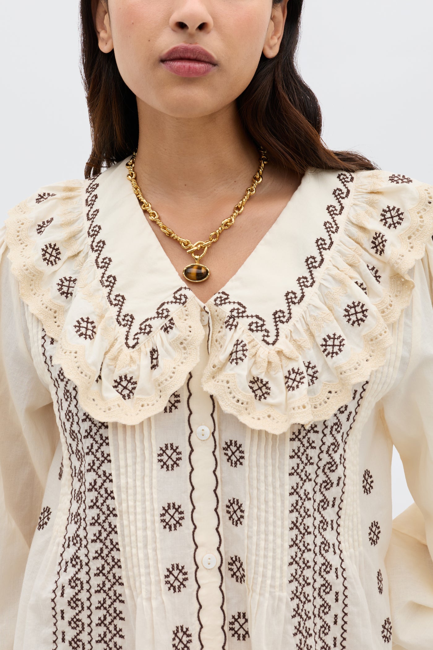 A woman wears the Percy - Mila Embroidery Brown cream blouse with a large ruffled lace collar, brown embroidery, and a gold oval pendant necklace. The image is cropped from her nose down to her chest.