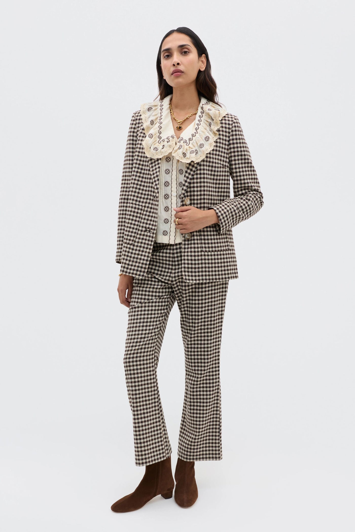 A woman stands against a plain light background, wearing the Percy - Mila Embroidery Brown suit with flared pants and a matching blazer, paired with a cream blouse with a wide frilled collar and brown ankle boots.