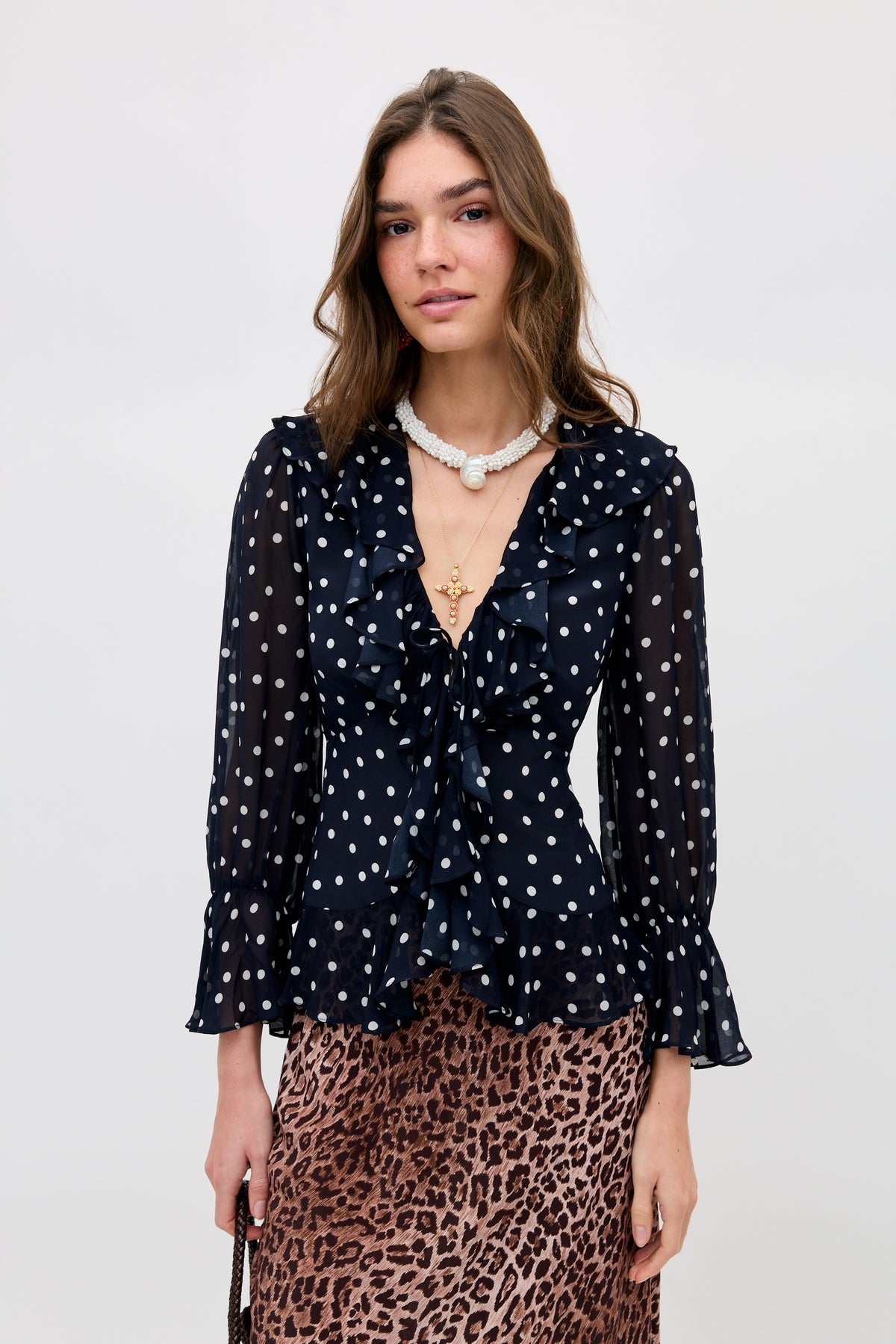 A woman poses against a light background wearing the Ami - Layla Dot Navy blouse, featuring sheer navy fabric with white polka dots, ruffled accents, and flared sleeves. She pairs it with a leopard print skirt and layered necklaces.