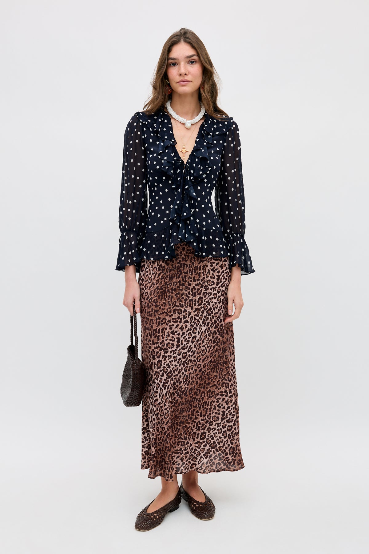 A woman stands against a plain background, wearing the Ami - Layla Dot Navy blouse with white polka dots, a brown leopard print maxi skirt, brown flats, a beaded necklace, and holding a small brown textured handbag.